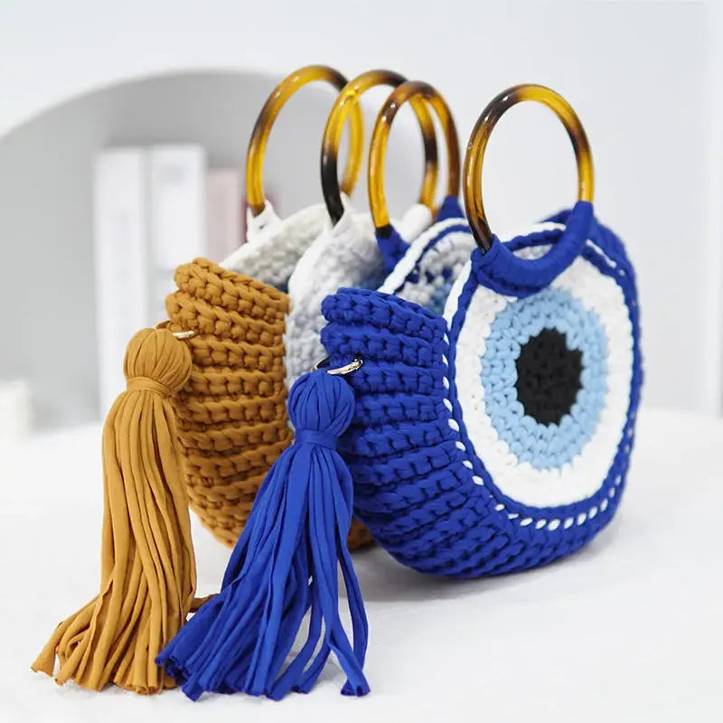 Duas bolsas Olho Grego em crochê sintético, nas cores azul e mostarda, com alças plásticas e detalhe do amuleto.
